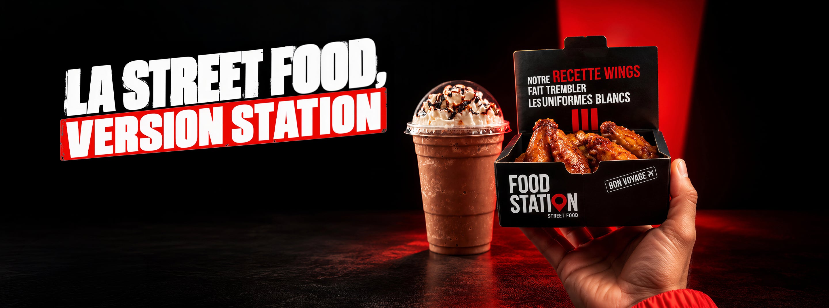 La street food, version station. Une main tend une boîte de wings Food Station, un milkshake au chocolat à côté.